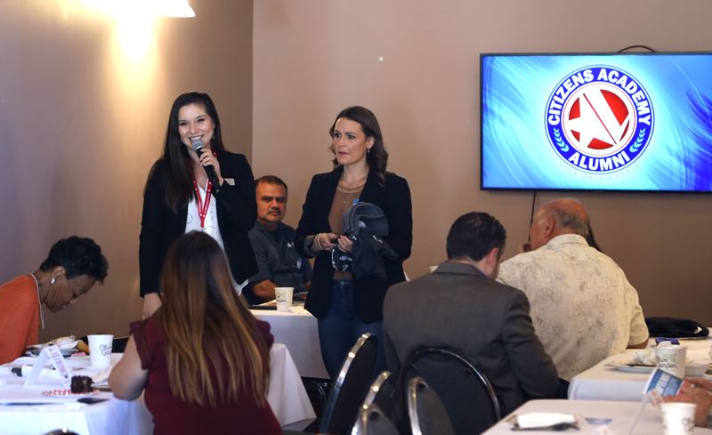 A woman standing near the front of some rectangle tables holds a microphone and smiles.