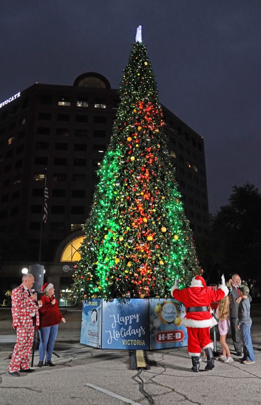 Santa Claus and a few other people gather at the base of a giant lighted Christmas tree