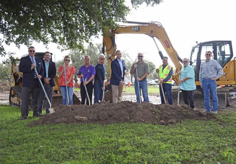 Group of people dig shovels into a pile of dirt with heavy machinery in the background