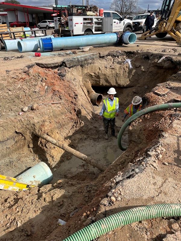 Two people in construction gear stand in a deep hole with the end of a pipe poking out of the side