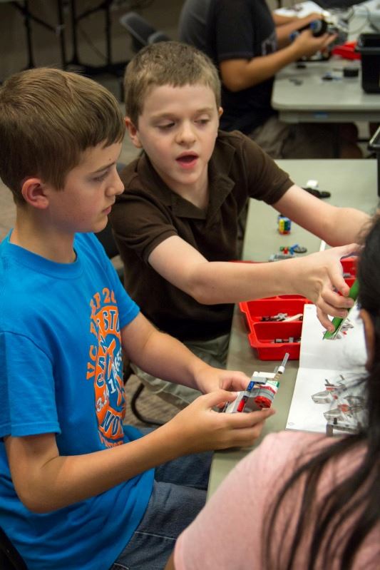 Two young children sit at a table constructing an item while reading the instructions together