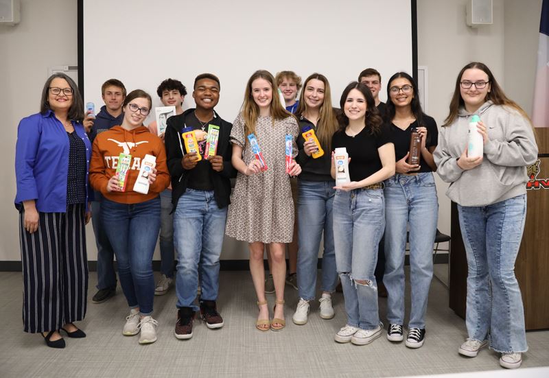 Students hold toothbrushes, shampoo, body wash and other hygiene items in group photo with a woman