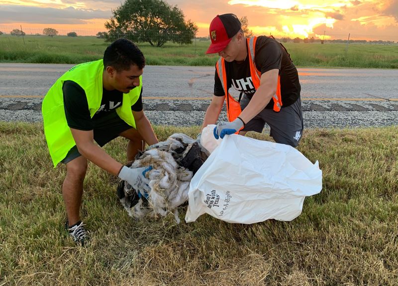 2 young men in work vests put a bundle of fabric in a trash bag on the side of the road