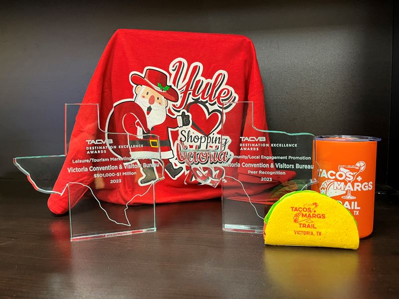 Two glass trophies, with a Yule Love Shopping shirt and Tacos and Margs tumbler and taco stress toy.