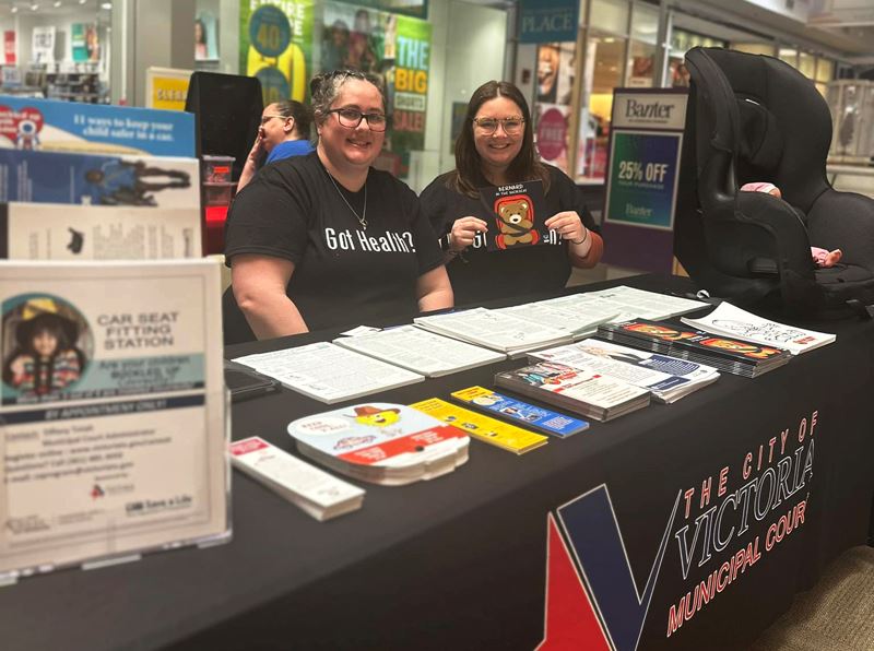 Two women pose at a Municipal Court booth covered in flyers and brochures at the Victoria Mall