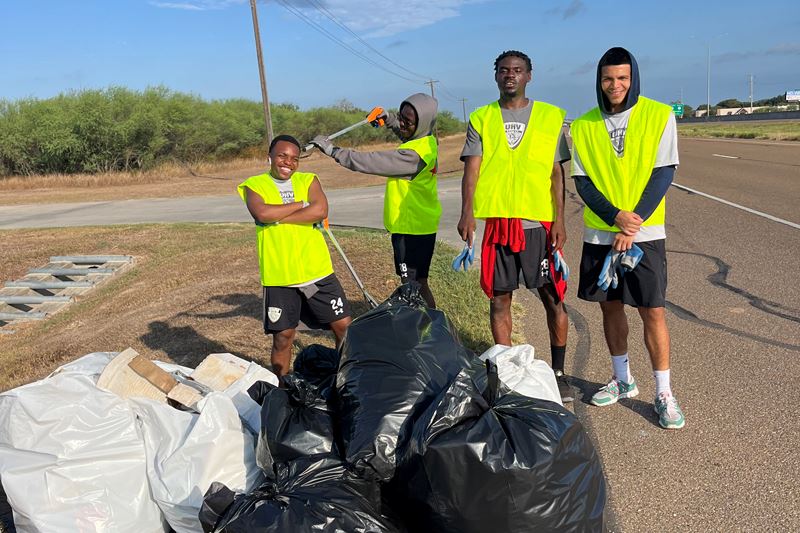 Three young men pose with trash grabbers near a pile of trash bags
