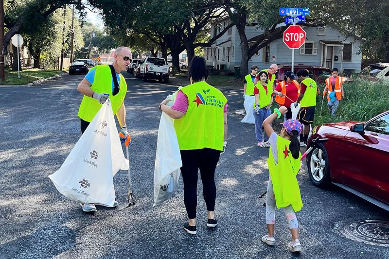 A group of children and adults walk through neighborhood holding trash bags and wearing safety vests