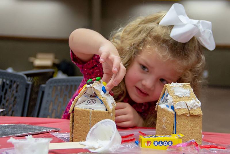 A young girl wearing a white hair bow carefully places M&Ms on a graham cracker gingerbread house.