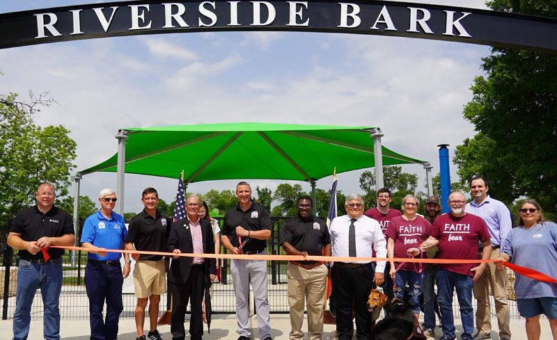 Group photo with ribbon in front of dog park under Riverside Bark sign