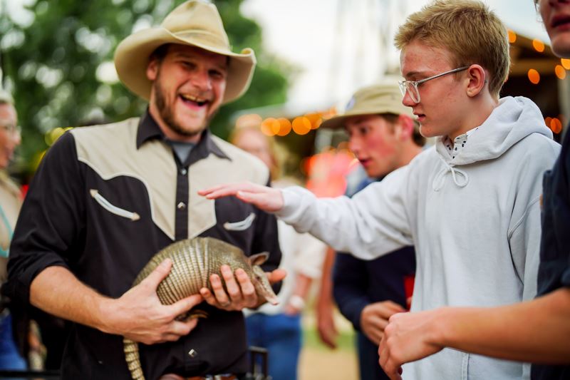 A young person tentatively reaches out to pet an armadillo held by a man in a cowboy hat