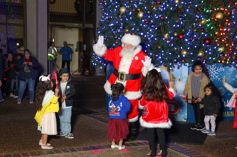 Santa Claus waving to children at the base of a giant lighted Christmas tree