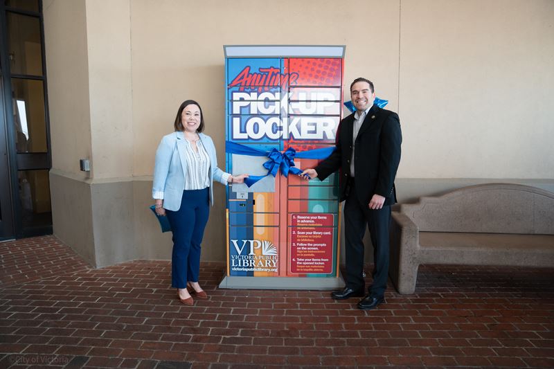 A woman and a man hold either end of a blue ribbon wrapped around the VPL locker at the mall