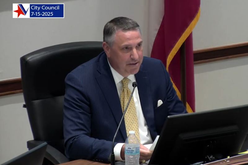Mayor Duane Crocker sitting at a council dais and reading into a microphone from printed notes
