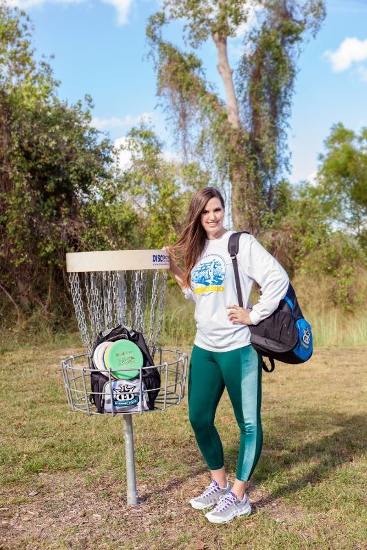 Smiling woman stands near a disc golf goal basket in Riverside Park that contains discs and a bag.