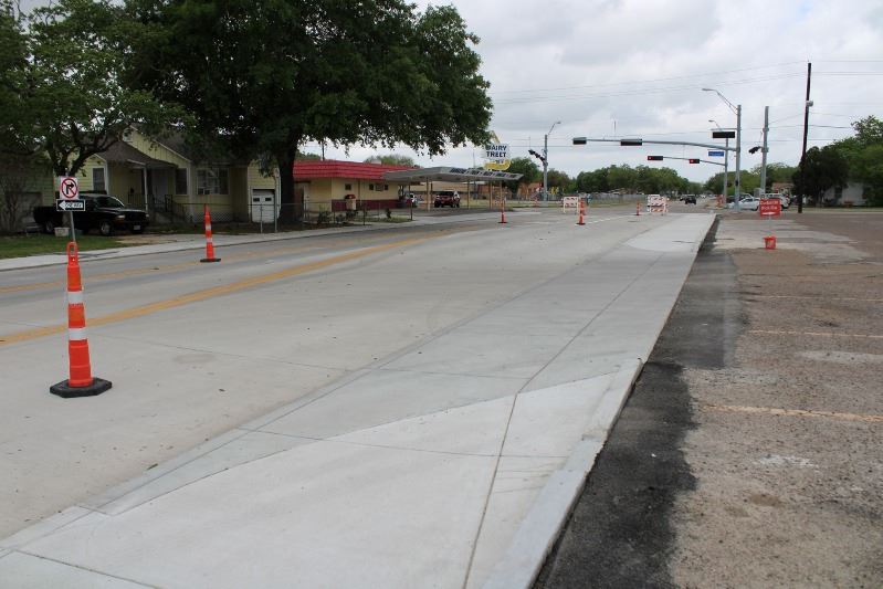 A concrete road bordered by a new sidewalk. The road is marked with cones and barricades.