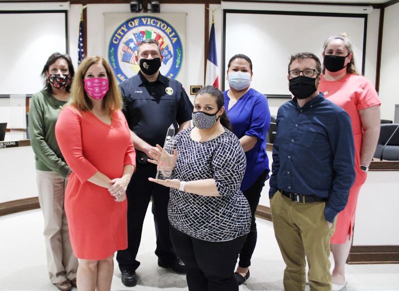 Seven people pose for group photo in the municipal courtroom. Woman in center holds a glass award.