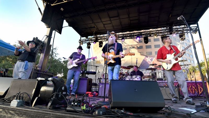 5 musicians perform onstage at DeLeon PLaza. 3 have guitars, 1 on drums, 1 holding a tambourine