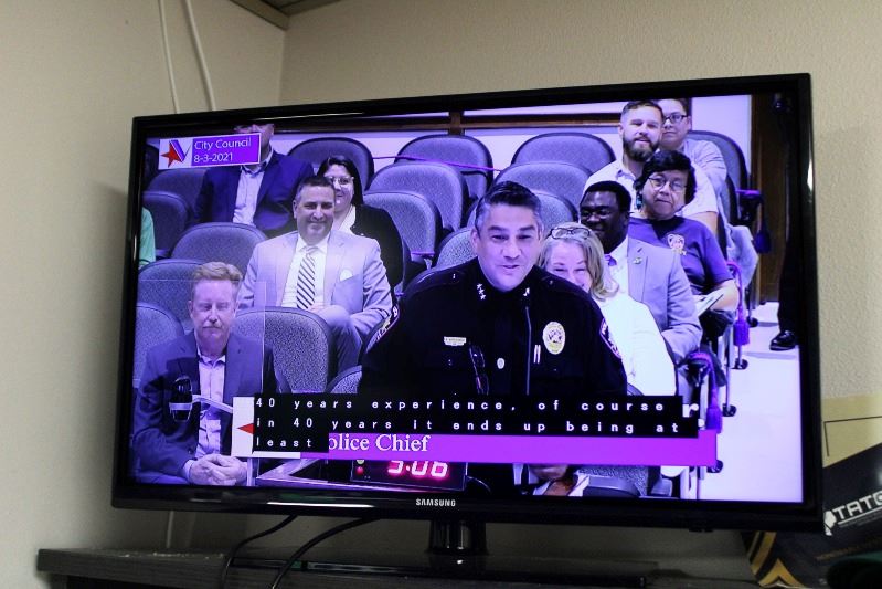 On TV screen, police chief speaks at City Council podium. Closed captioning is displayed.