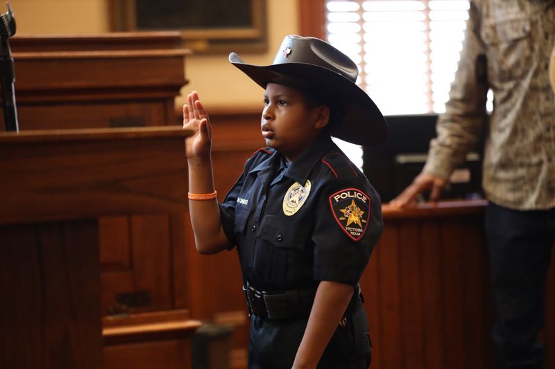 A child in police uniform raises his hand during a swearing-in ceremony at a courthouse.