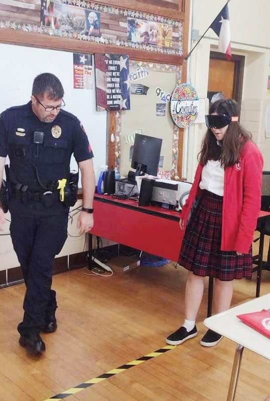 A uniformed police officer helps a girl wearing impairment goggles walk along a line