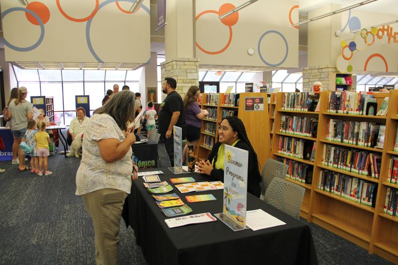 A woman sits at a table with signs reading Summer Programs for Adults. Another woman looks at flyers