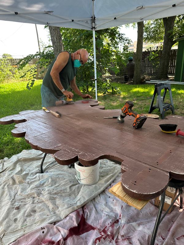 Sean Watkins uses a handheld sander to sand the back of a sign under a canopy outdoors