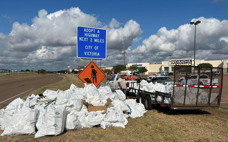 Bags of trash are piled on the ground and in a trailer by an Adopt a Highway sign