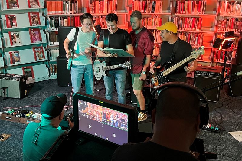 Two men use video equipment to film a group of men holding guitars in a library