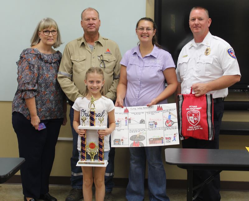 A girl holding a trophy. Four adults stand behind her holding a poster and a red bag.