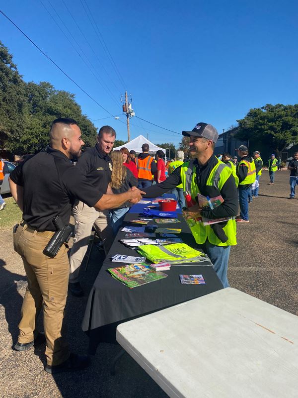 Two people in police department polos stand at an outdoor table and greet a man in a safety vest
