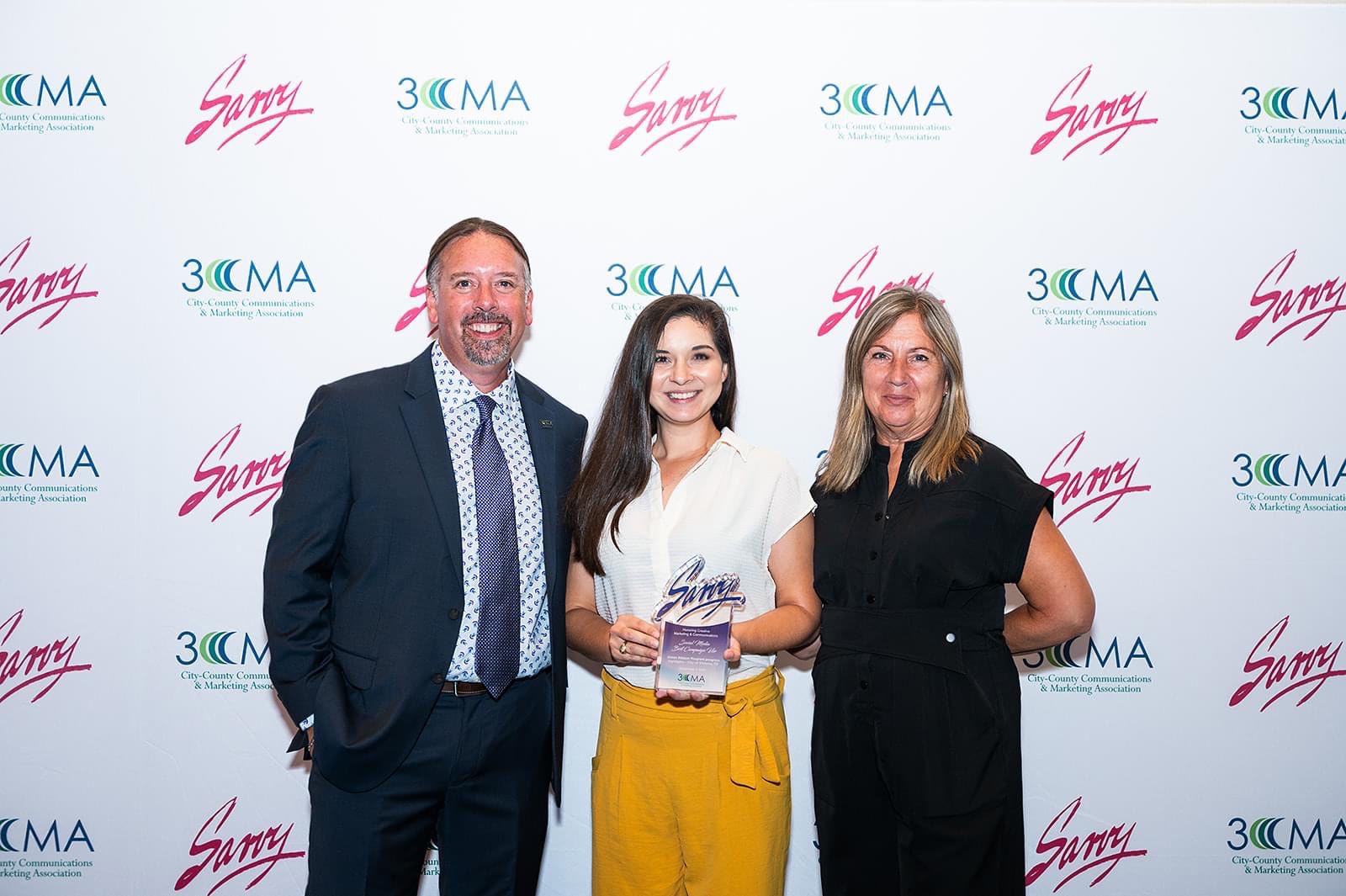 Three people standing behind a white backdrop. Lady in the middle is holding an award.