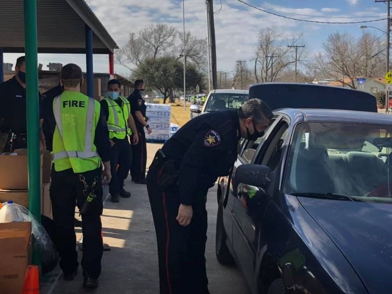 Workers in Police and Fire uniforms assist at a drive-thru event. Cars are lined up down the street.