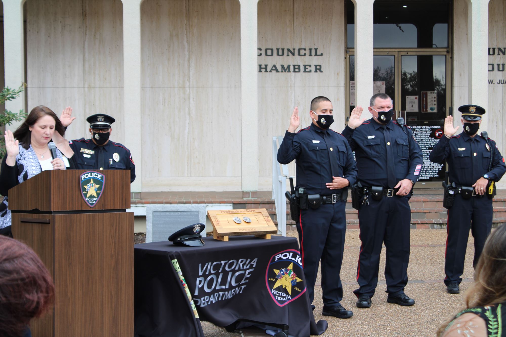 Woman at outdoor podium raises hand. Uniformed police officers raise their hands.