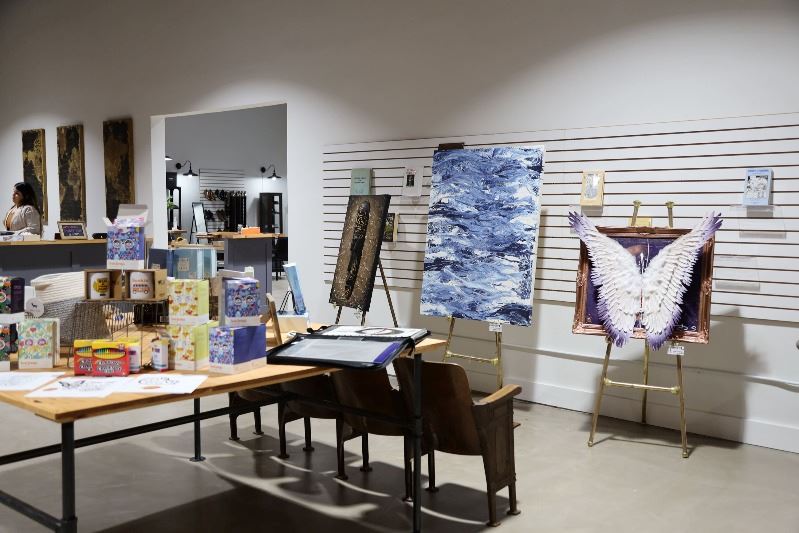 Paintings, some with 3D components, sit on easels at Texian Books. Merchandise sits on table.