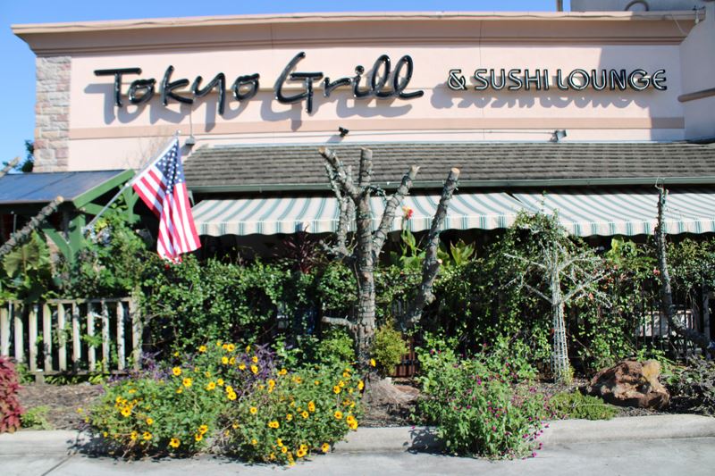 Lighted trees and flowering plants fill a bed along a covered patio at Tokyo Grill. American flag.