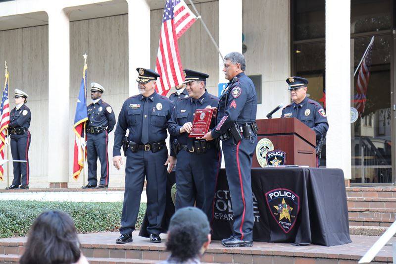 Three uniformed police officers stand at a makeshift stage at the police station. One holds a plaque