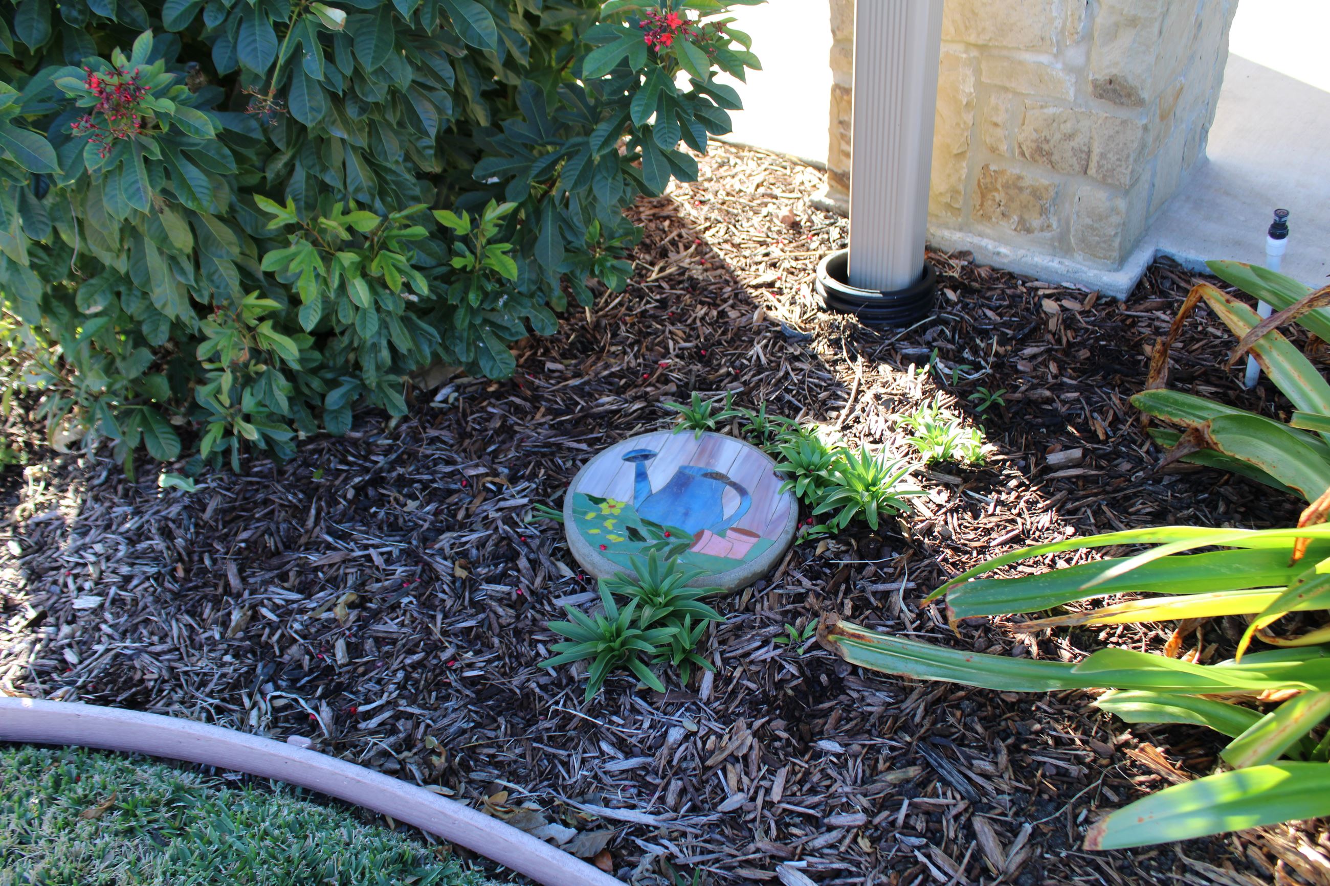 A decorative stepping-stone with an image of a watering can sits in a flowerbed