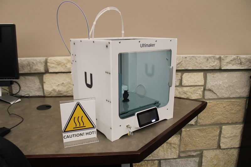 A cube shaped Ultimaker brand 3D printer. Sign reads Caution, Hot