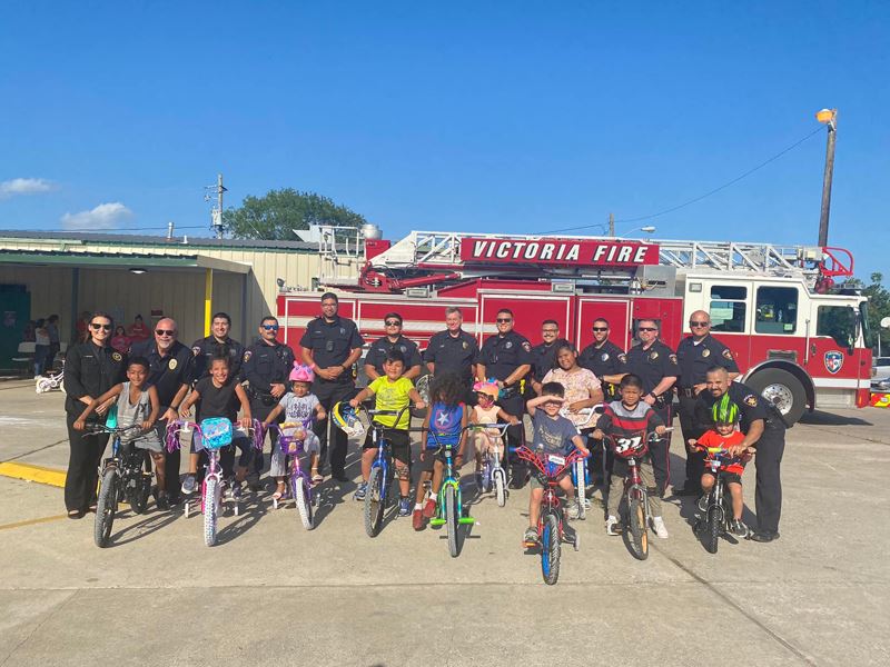 Nine kids on bikes pose with uniformed police officers in front of a fire truck at Christ's Kitch